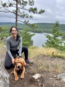 A woman smiling with her dog among the trees overlooking a lake.